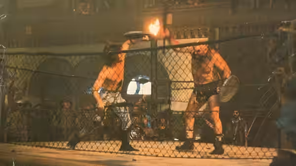 Men face off inside a chain-link cage as a fire burns behind them