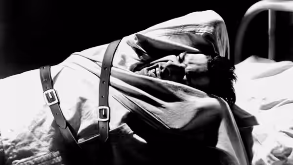 A man lies strapped to a hospital bed, shielding his face with an arm