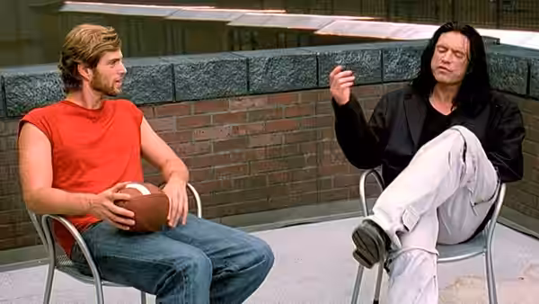 Johnny (Tommy Wiseau) gesturing to Mark (Greg Sestero) while seated on the rooftop