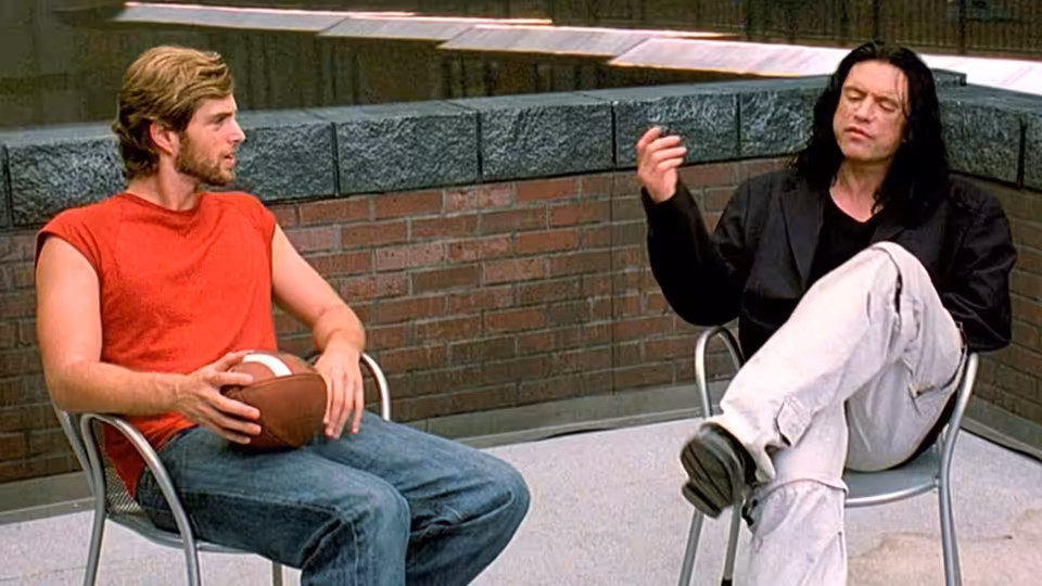 Johnny (Tommy Wiseau) gesturing to Mark (Greg Sestero) while seated on the rooftop