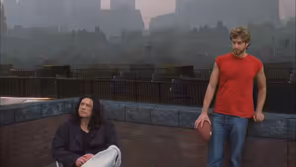 Mark (Greg Sestero) standing with football on rooftop near seated Johnny (Tommy Wiseau)