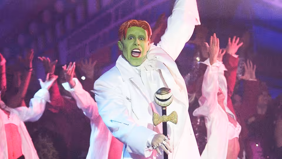 Loki (Alan Cumming) sings on stage with arm raised and crowd cheering