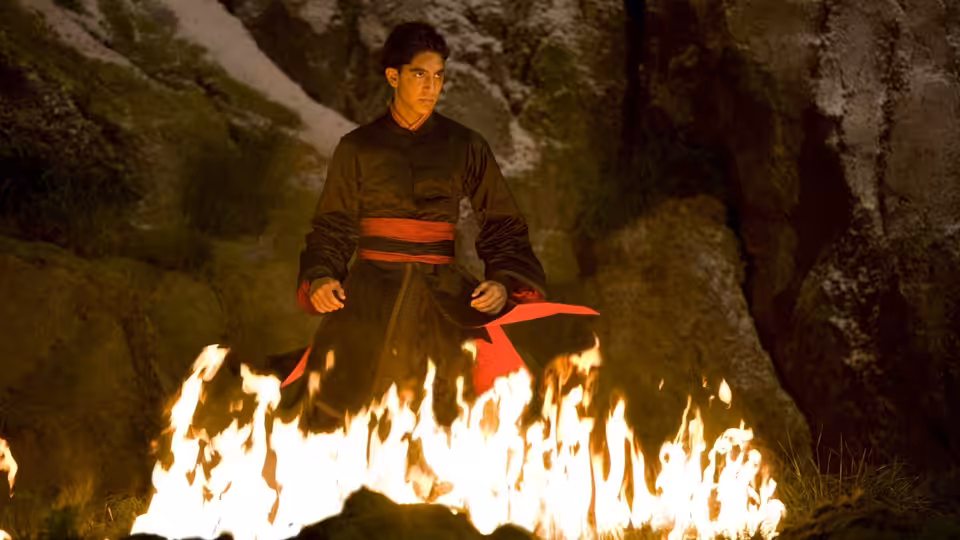 Prince Zuko (Dev Patel) seated before a large fire in rocky terrain