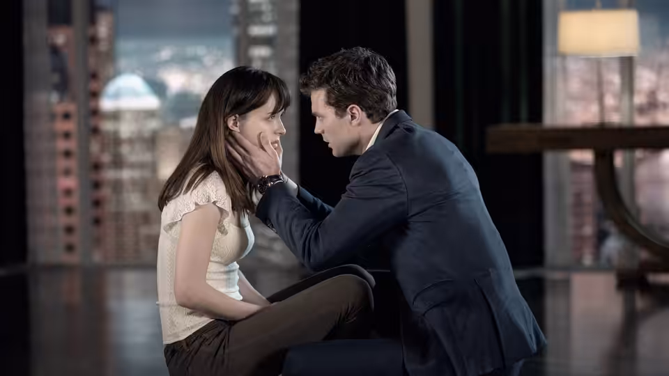 Christian Grey (Jamie Dornan) touching Anastasia Steele (Dakota Johnson) face while seated indoors