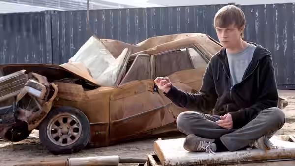 Andrew Detmer sits cross-legged beside a wrecked car, lifting one hand