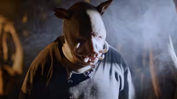 A bloodied pig-masked man stands amid smoke in a dark outdoor setting