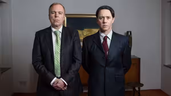 Two suited men standing side by side in a plain office interior
