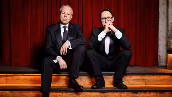 Two men in black suits seated on wooden steps before a red curtain