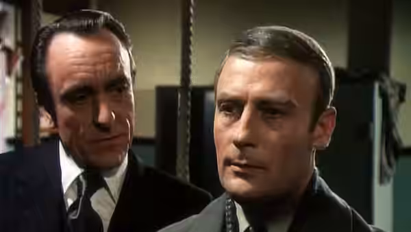 Two men in dark suits stand together looking ahead intently