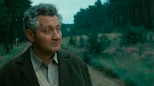 Man with curly gray hair stands on dirt path amid trees and fields