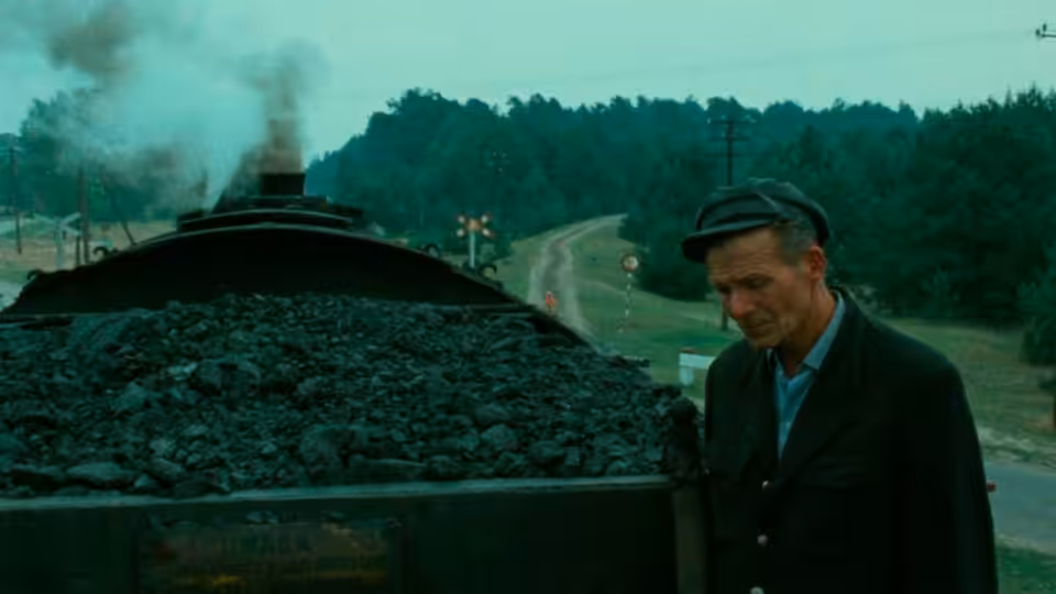 Man in cap stands beside coal-filled train car on rural railway tracks