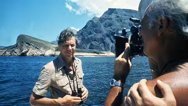 David Attenborough holding binoculars on a boat as a cameraman films him