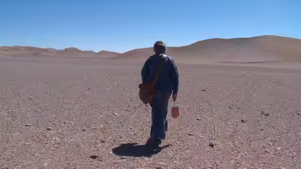 A person walks across a barren desert carrying a bag and container