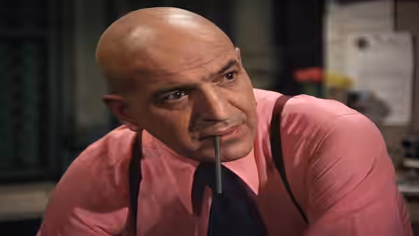 Kojak (Telly Savalas) leans forward with a pencil between his lips