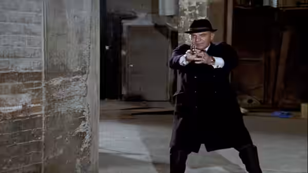 Kojak (Telly Savalas) stands in a warehouse aiming a handgun with both hands