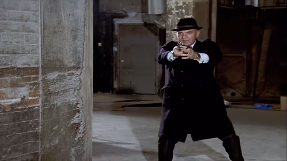 Kojak (Telly Savalas) stands in a warehouse aiming a handgun with both hands