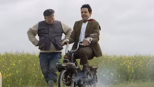 Mr. Bean (Rowan Atkinson) rides a small motorcycle beside a man in a field