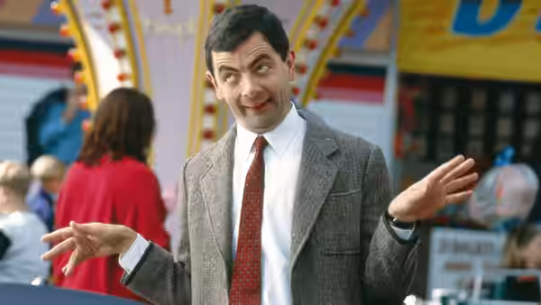 Mr. Bean (Rowan Atkinson) raises both hands at a brightly decorated fairground stall