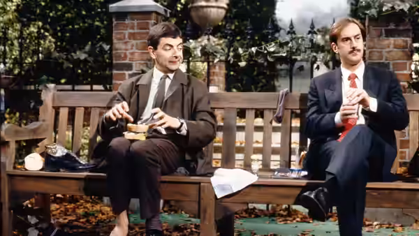 Mr. Bean (Rowan Atkinson) and a suited man eat lunch on a park bench