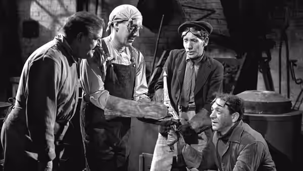 Four men examine a small object in a cluttered workshop