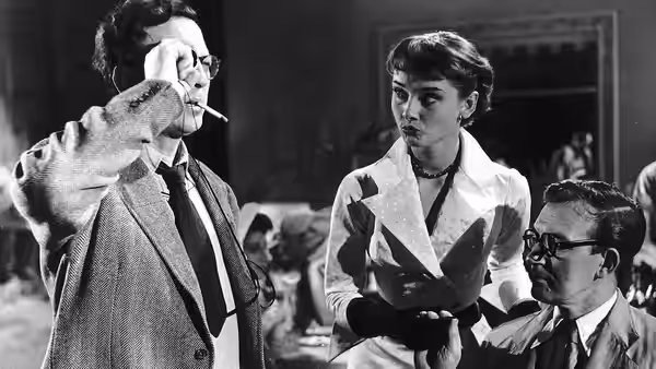 A woman stands between a man smoking and a seated bespectacled man
