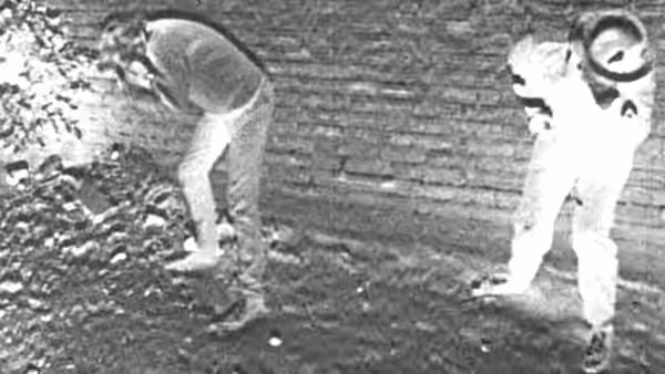 One person bends over debris while another stands beside a brick wall