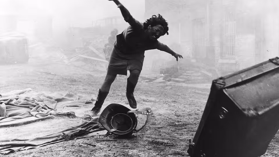 A person staggers with arms outstretched among debris in a devastated street