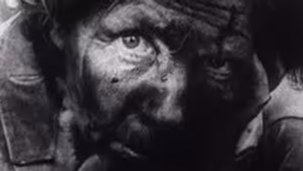 A weary man in a helmet looks ahead from beneath a dark hood