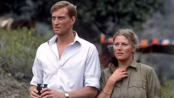 Man holding binoculars stands beside woman with hand on her chest outdoors