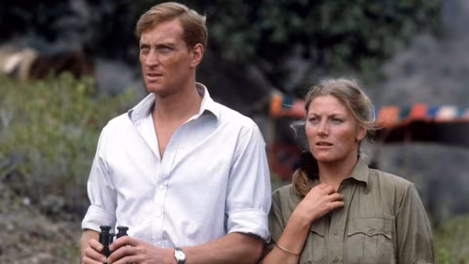 Man holding binoculars stands beside woman with hand on her chest outdoors