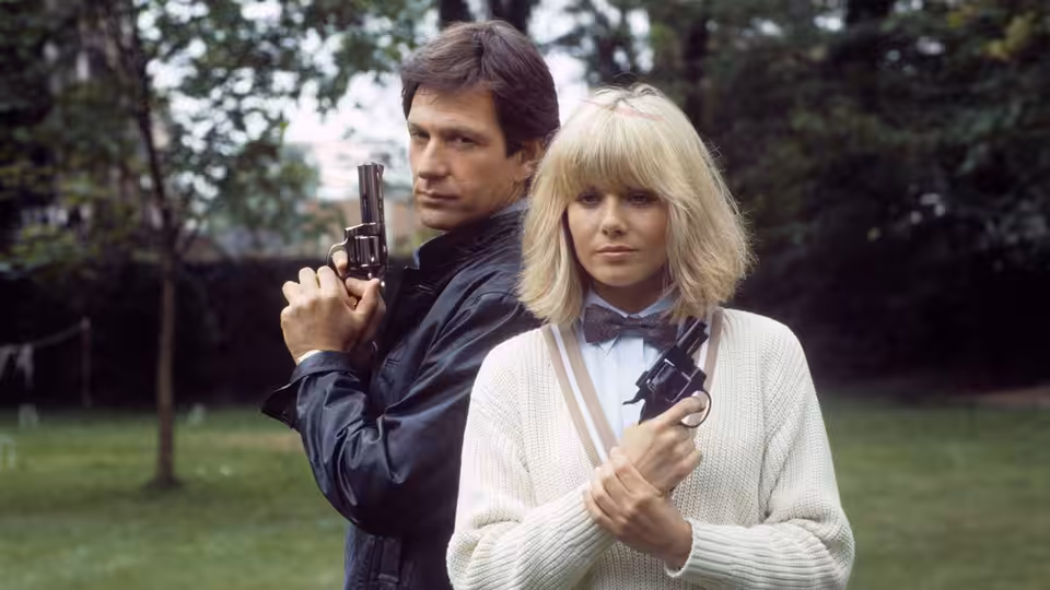 Dempsey (Michael Brandon) and Makepeace (Glynis Barber) holding guns in a garden