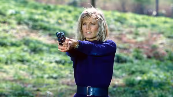 Makepeace (Glynis Barber) aiming a revolver in a grassy field
