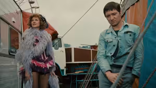 A red-haired performer in a feathered costume stands beside a man near a circus truck