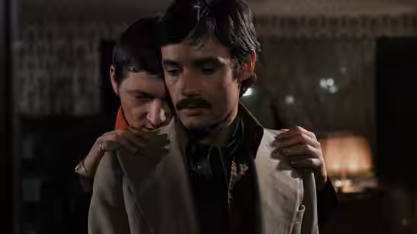 A man embraces another from behind in a dimly lit room
