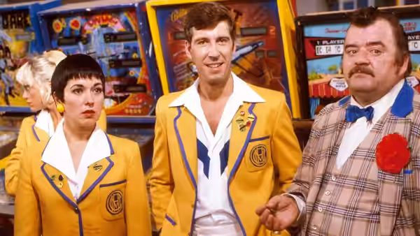 Woman with short dark hair in yellow jacket stands with two men near arcade machines