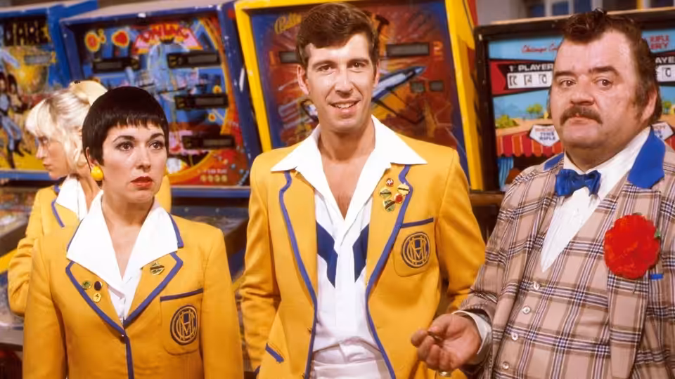 Woman with short dark hair in yellow jacket stands with two men near arcade machines