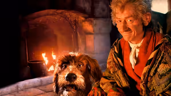 The Storyteller (John Hurt) smiles at his dog beside a stone fireplace