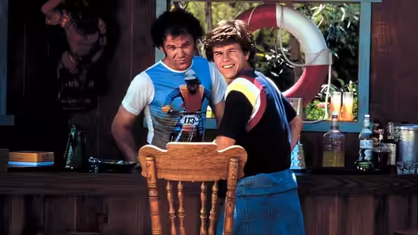 two men standing at a wooden bar one leaning on a chair back smiling