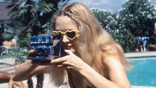 a blonde woman holding a Polaroid camera to her eye by a pool