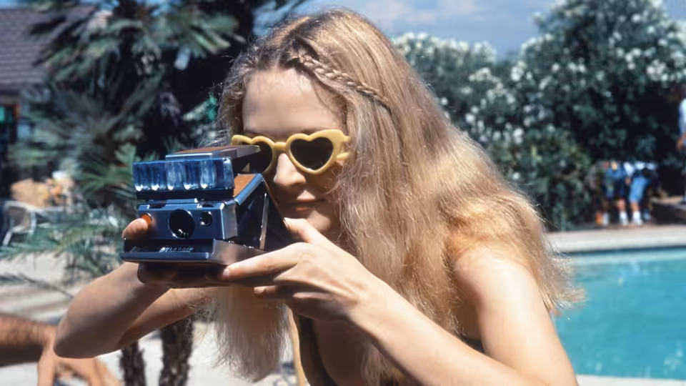 a blonde woman holding a Polaroid camera to her eye by a pool
