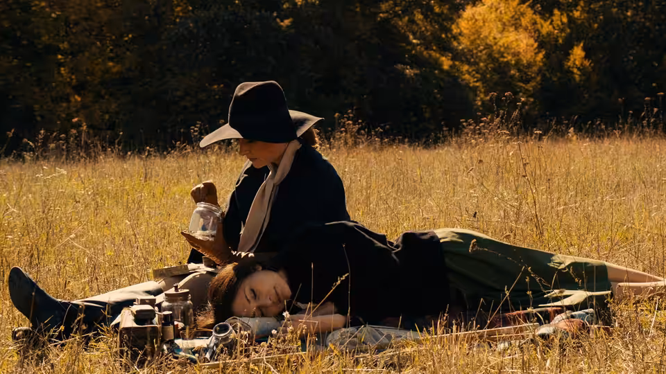 Woman in dark hat sits in meadow beside reclining woman during picnic