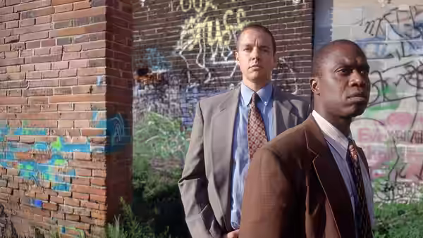 Two suited men stand in front of a graffiti-covered brick wall