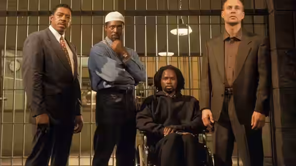 Four men stand before prison bars with one seated in a wheelchair