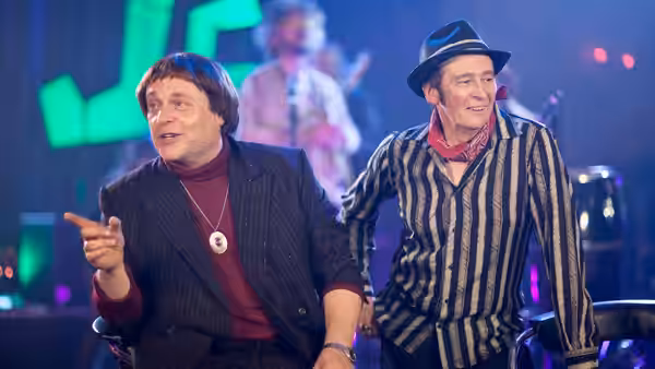 Two men stand on a stage, one pointing forward while the other smiles
