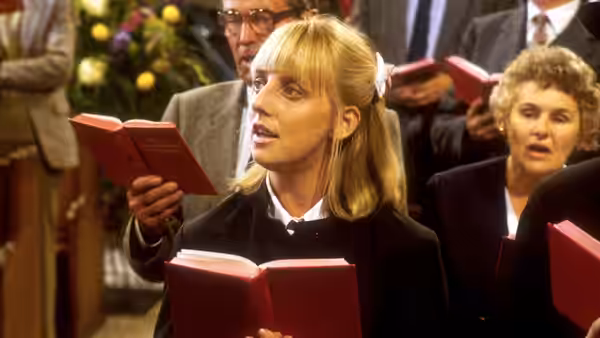 Alice (Emma Chambers) singing from a red hymn book during a service