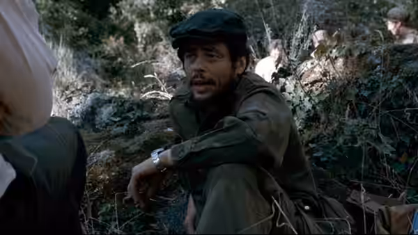 Che Guevara (Benicio del Toro) seated on the ground in a forest clearing