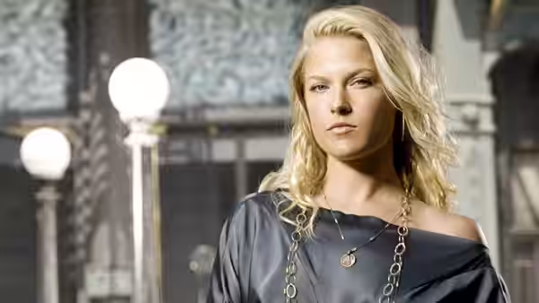 A blonde woman in a dark off-shoulder top stands in a grand interior