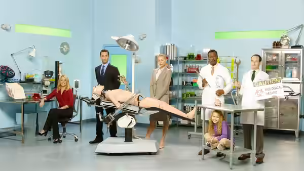 Professionals in lab coats and suits stand around a dummy on an exam table in a bright laboratory