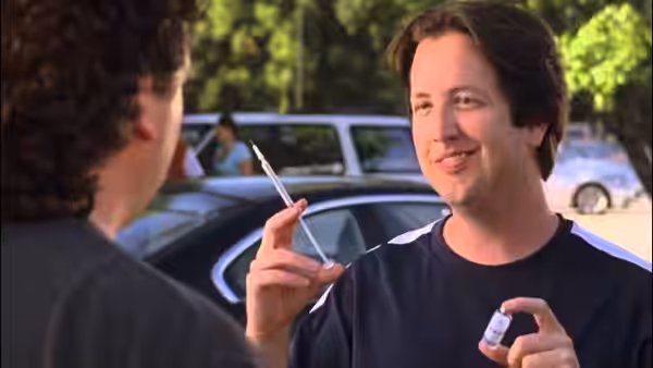A man holds a syringe and small bottle while facing another man near parked cars