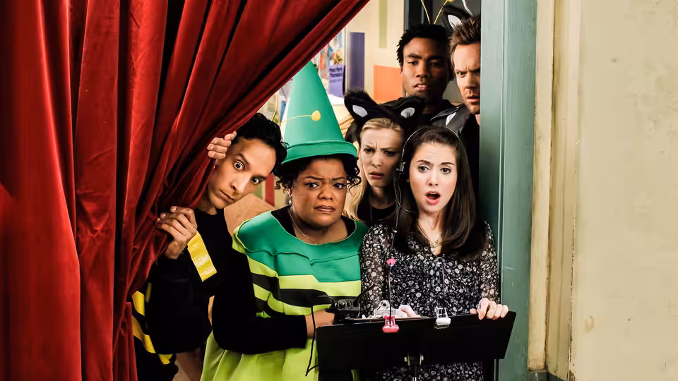 Annie (Alison Brie) holds a tray while costumed friends peer from behind a red curtain